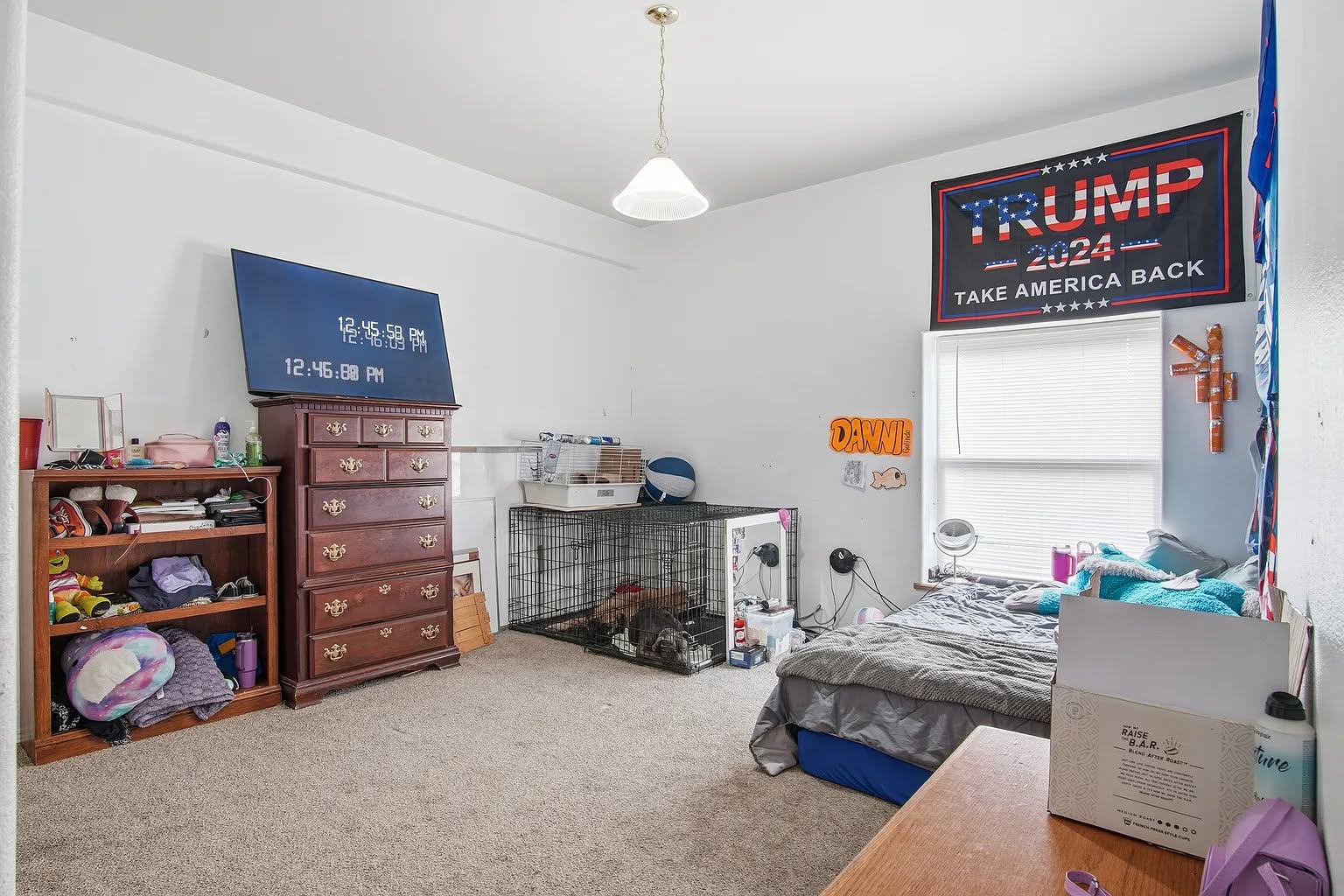 A bedroom with a wooden dresser and shelves, a wall TV, and a dog crate. A bed is near the window, and a large 'Trump 2024' flag hangs above it. The room is tidy and spacious.