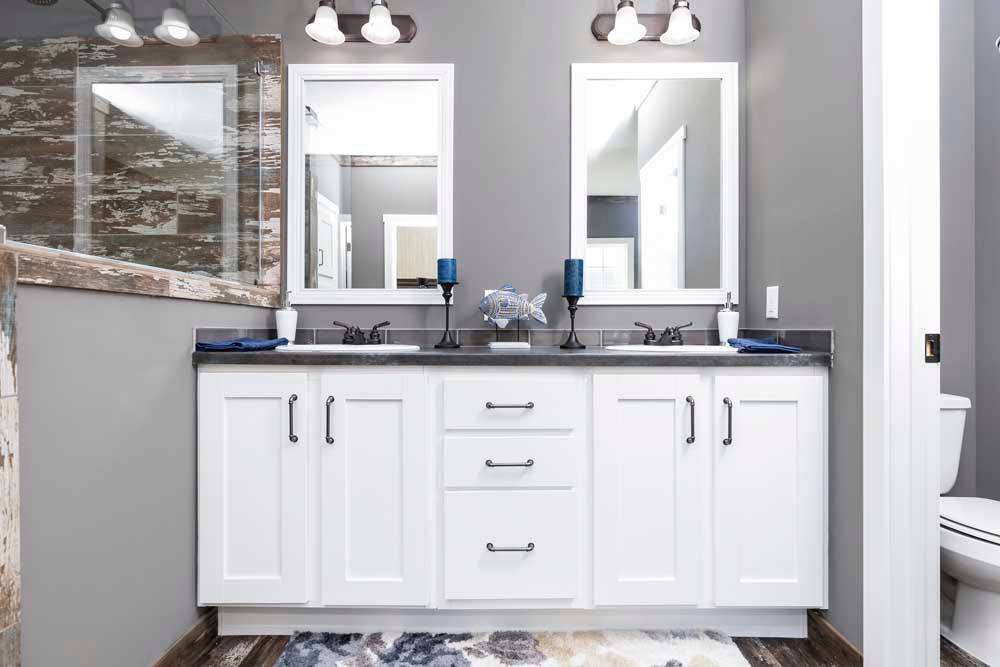 Modern bathroom with dual sinks, white cabinets, and black countertops. Two mirrors are above, with blue candle holders between them. Gray walls and warm lighting create a serene atmosphere.
