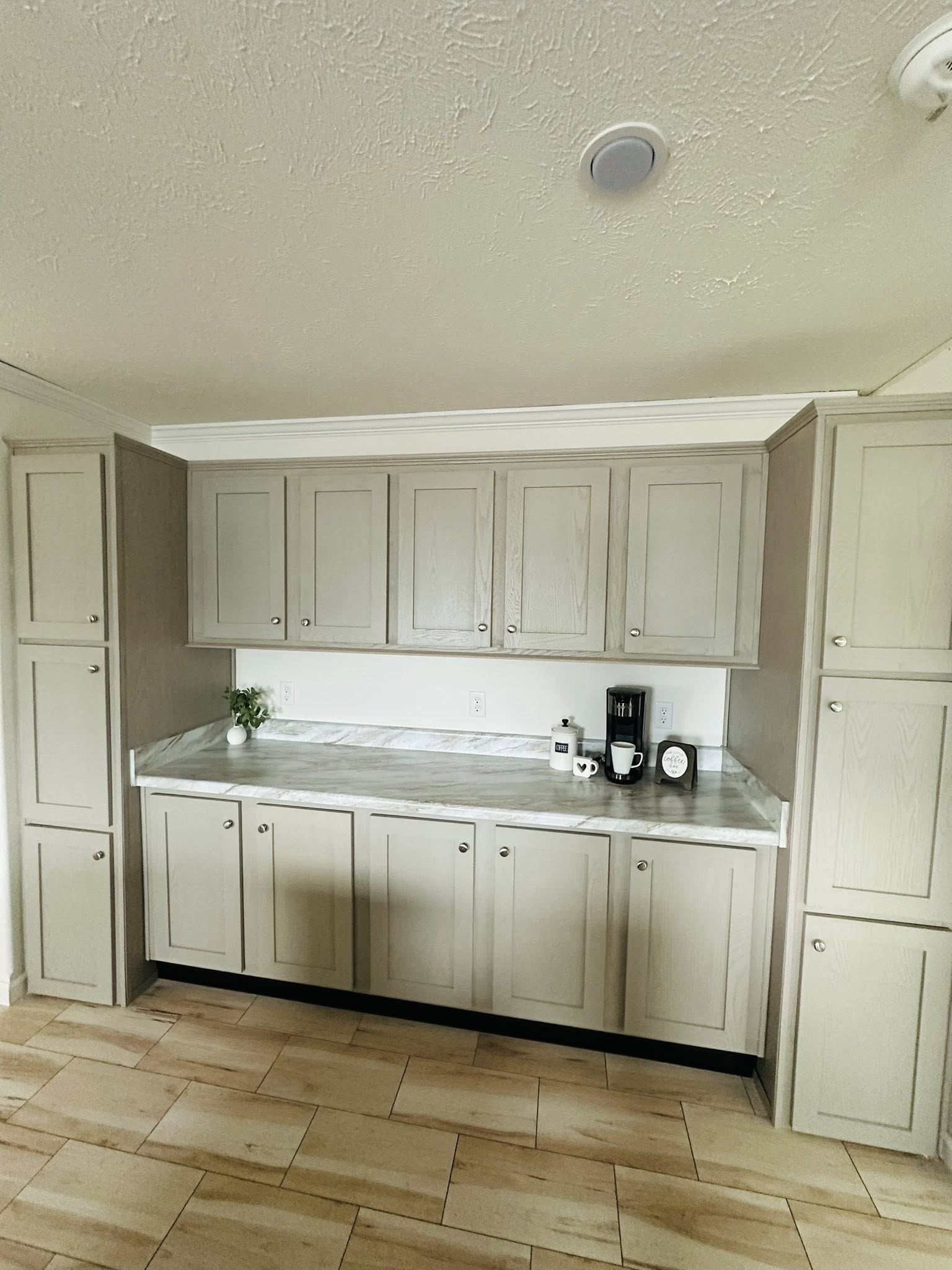 Minimalist kitchen with light gray cabinets, marble countertops, and beige tiled floor. Features a coffee maker, mugs, and a small plant, conveying a clean, modern feel.