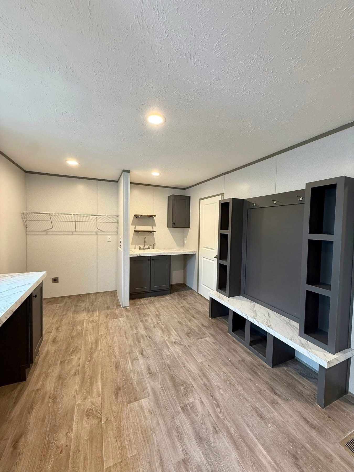 Modern utility room with wood flooring, white walls, and recessed lighting. Features dark cabinets, a small sink area, shelves, and a built-in bench.