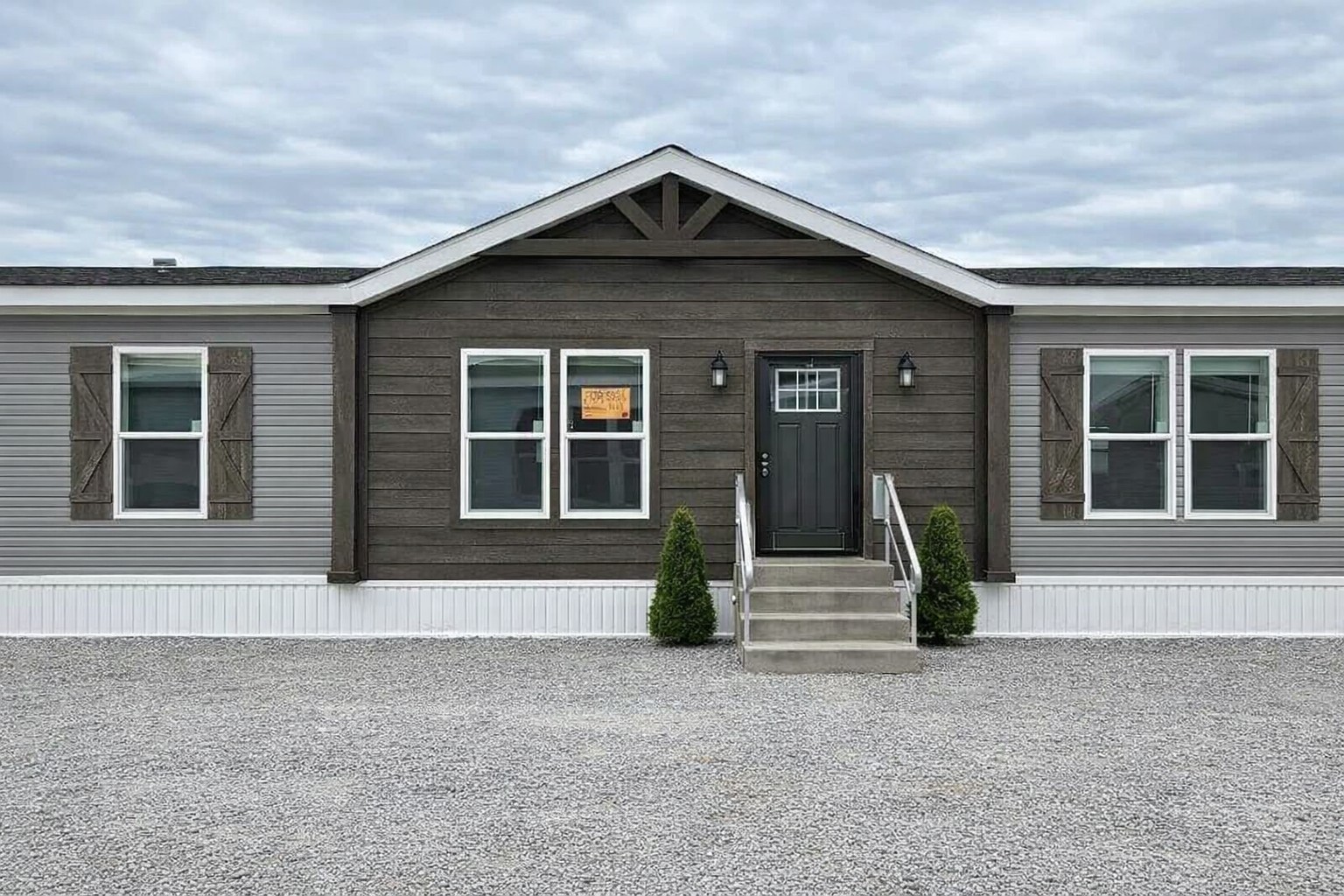 Single-story modular home with dark wood siding and a black front door, flanked by symmetrical windows with shutters. Two small shrubs line the entryway steps.