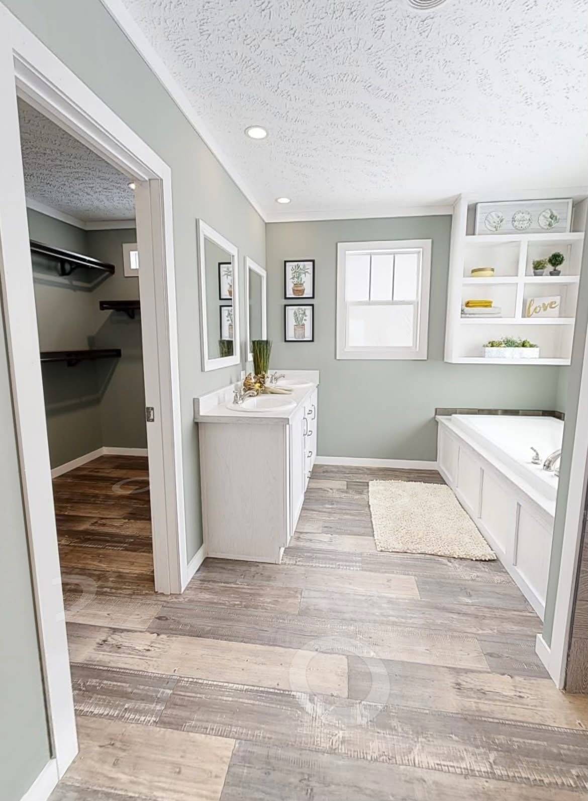 Bright bathroom with wood-style flooring, a white vanity, and decor on shelves. Soft green walls and art provide a calm, inviting atmosphere.
