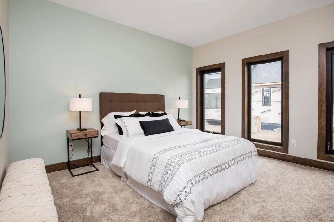 A serene bedroom features a neatly made bed with white and patterned linens, flanked by two lamps on wooden nightstands. Three large windows let in natural light, enhancing the calm ambiance.