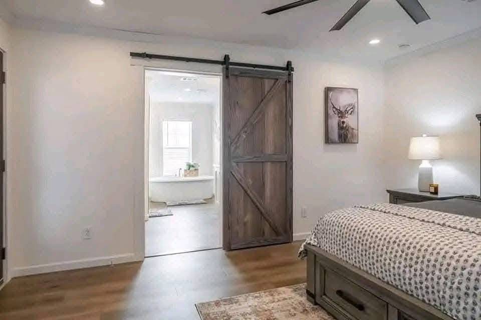 A cozy bedroom with a wooden sliding barn door leading to a bright bathroom. Features a soft-lit lamp, rustic deer artwork, and a patterned bedspread.