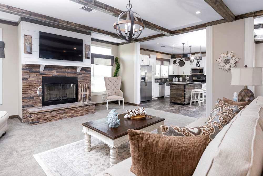 Spacious living room with beige sofas, patterned cushions, a wooden coffee table, and a stone fireplace. The room leads into a modern kitchen. Warm, welcoming ambiance.