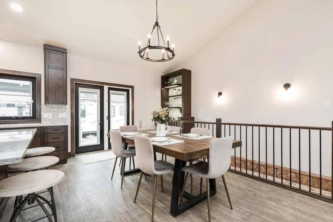 Modern dining room with light wood flooring, a wooden table set for four with beige chairs, black chandelier, and glass doors leading outside.