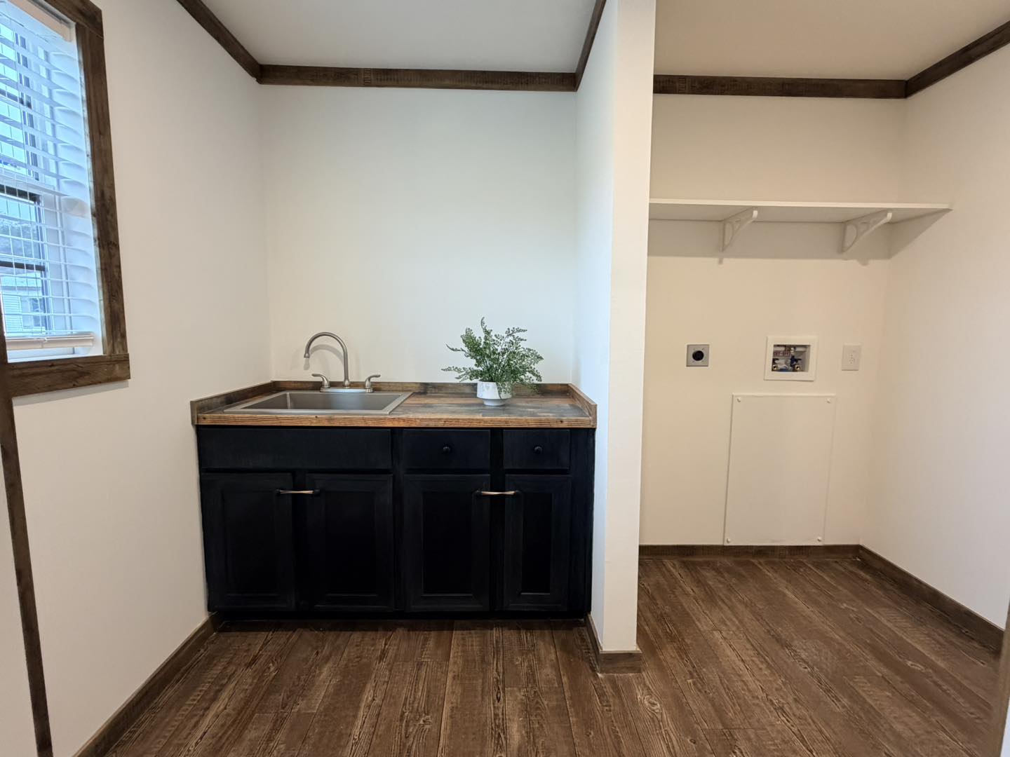 Laundry room with a dark wooden cabinet and sink, a small green plant on top. To the right, an open area for appliances with a shelf above. Bright and tidy space.