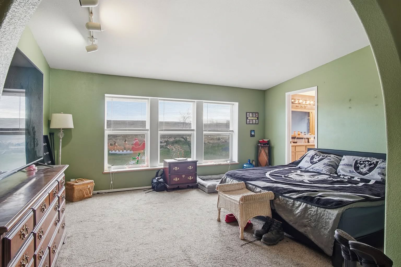 Spacious bedroom with green walls features a large bed, dresser, and wicker stool. Three windows provide ample light. Cozy and organized with a relaxed ambiance.