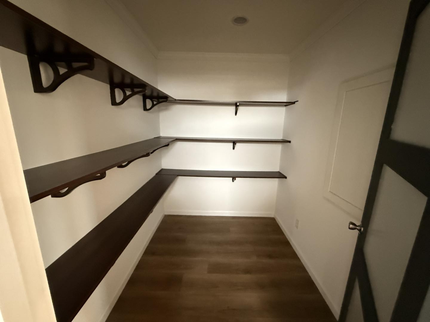 Empty pantry with dark wooden shelves lining white walls on three sides, brown wooden floor, and a single recessed light, creating a minimalist feeling.