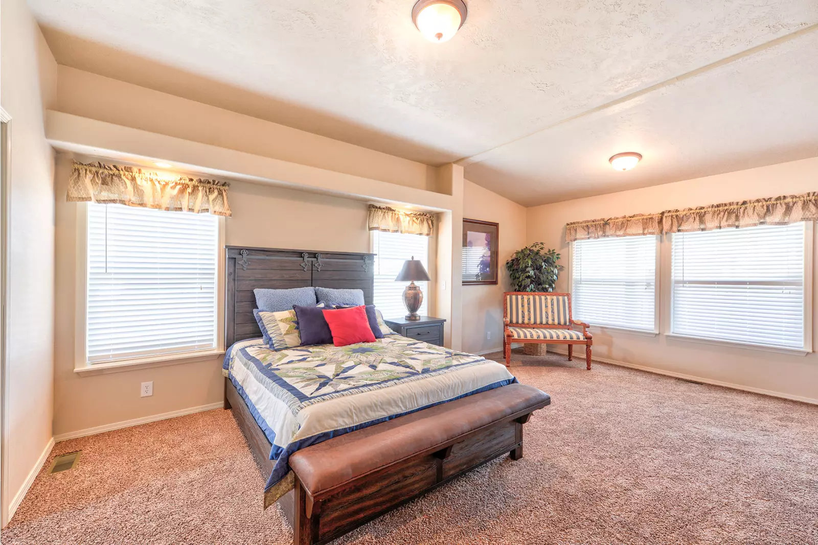 Spacious bedroom with a large bed featuring colorful cushions and a patterned quilt, flanked by two windows. A small bench and plant add warmth.