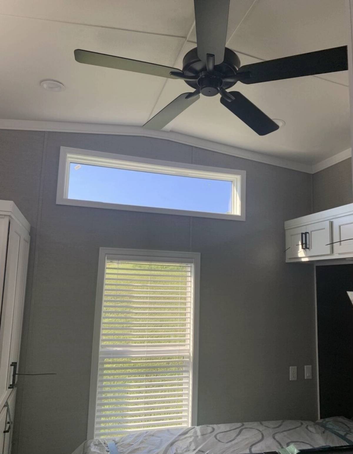 Ceiling fan with black blades in a room with gray walls. Two windows, one high and rectangular, the other vertical with blinds, provide natural light.