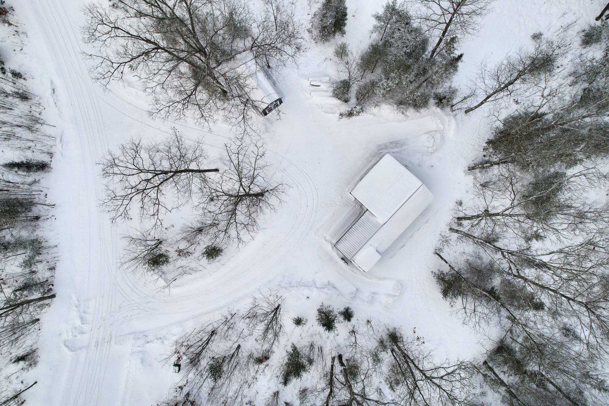 Aerial view of a snow-covered cabin surrounded by leafless trees in a forest. Snow trails weave through the scene, creating a serene, wintery atmosphere.