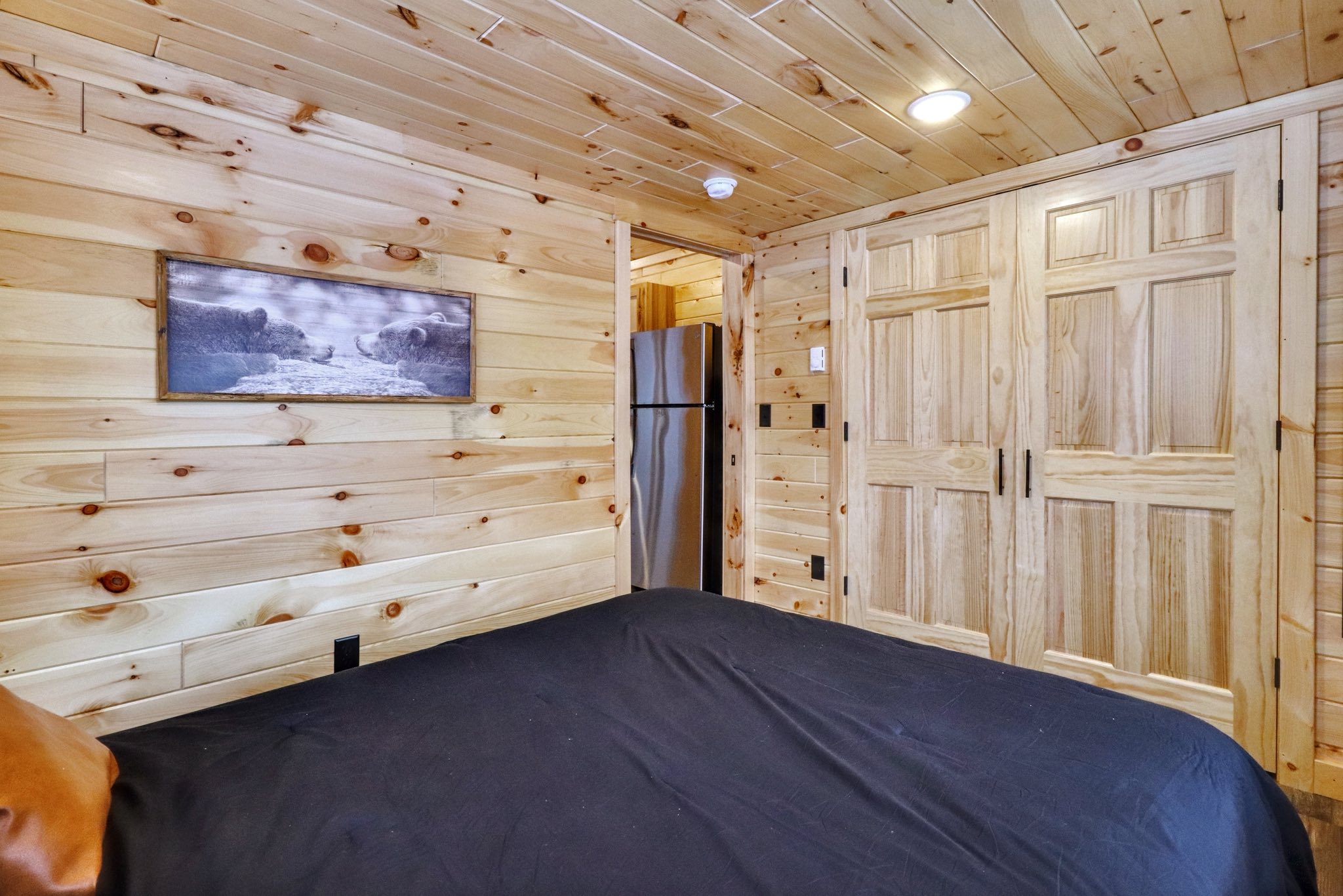 Cozy wooden cabin bedroom with a black bedspread and artwork on the wall. A fridge is visible through the door, and light wood paneling adds warmth.