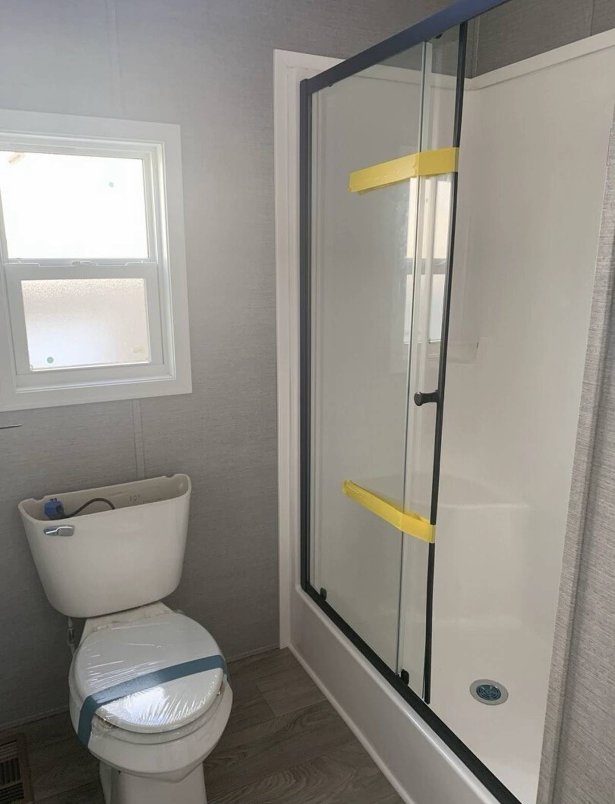 A small bathroom with a window, toilet covered in plastic, and a glass shower door with yellow tape. The setting is clean and minimalistic.