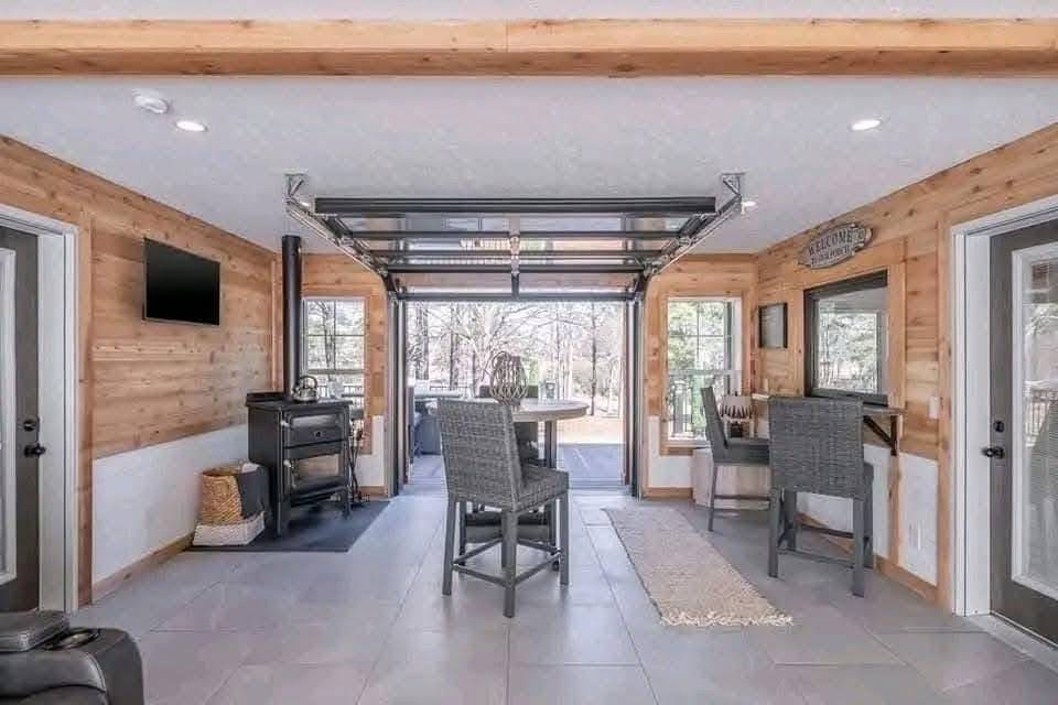 Cozy room with wood-paneled walls, large windows, and a glass garage door opening to a patio. Features a wood stove, bar-height table, and modern chairs.