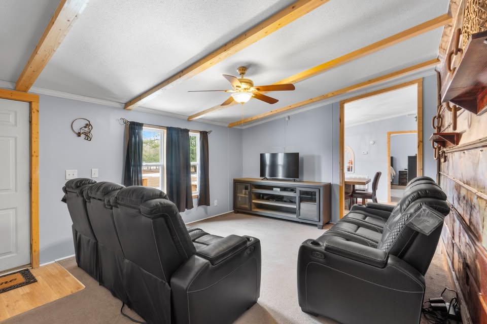 A cozy living room with four black recliners facing a large flat-screen TV on a wooden stand. Ceiling fan, wooden beams, gray walls. Sunlight through window.