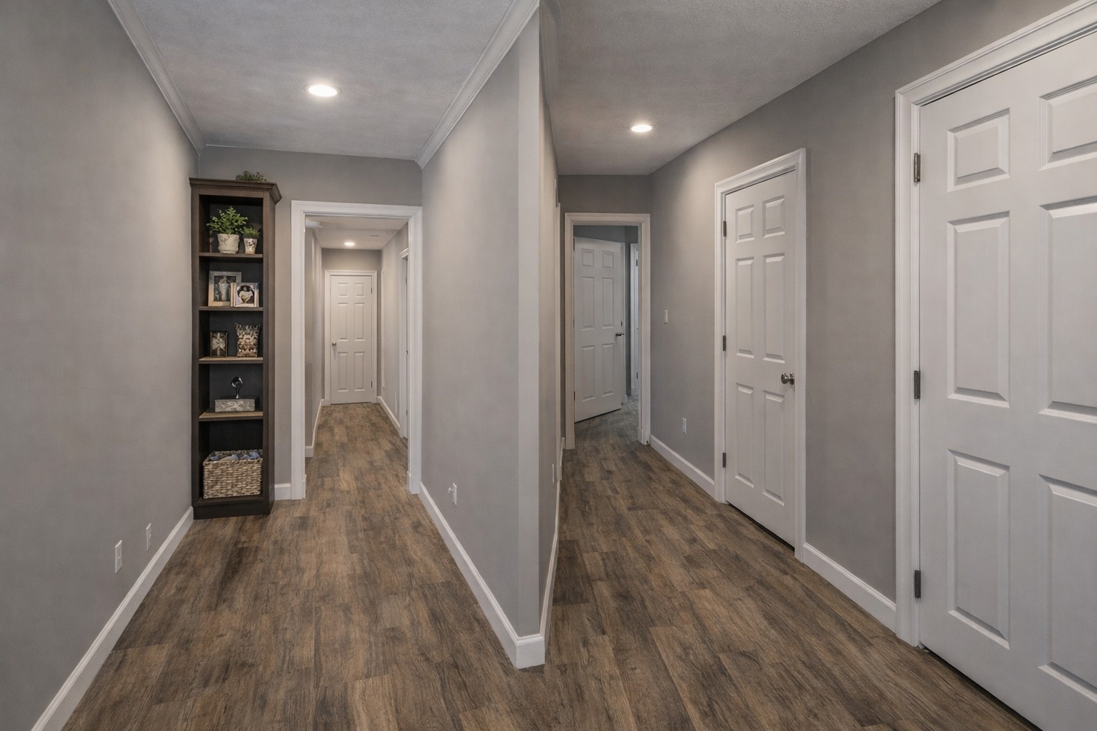A well-lit hallway with gray walls and wood flooring. The space features two doorways and a dark wood bookshelf filled with photos and decor. Calm ambiance.