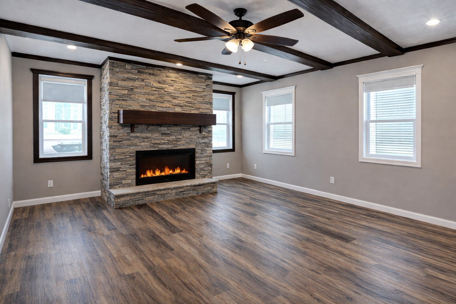 Spacious living room with wood flooring, a stone fireplace, and a ceiling fan. Large windows allow natural light. Modern, cozy ambiance.