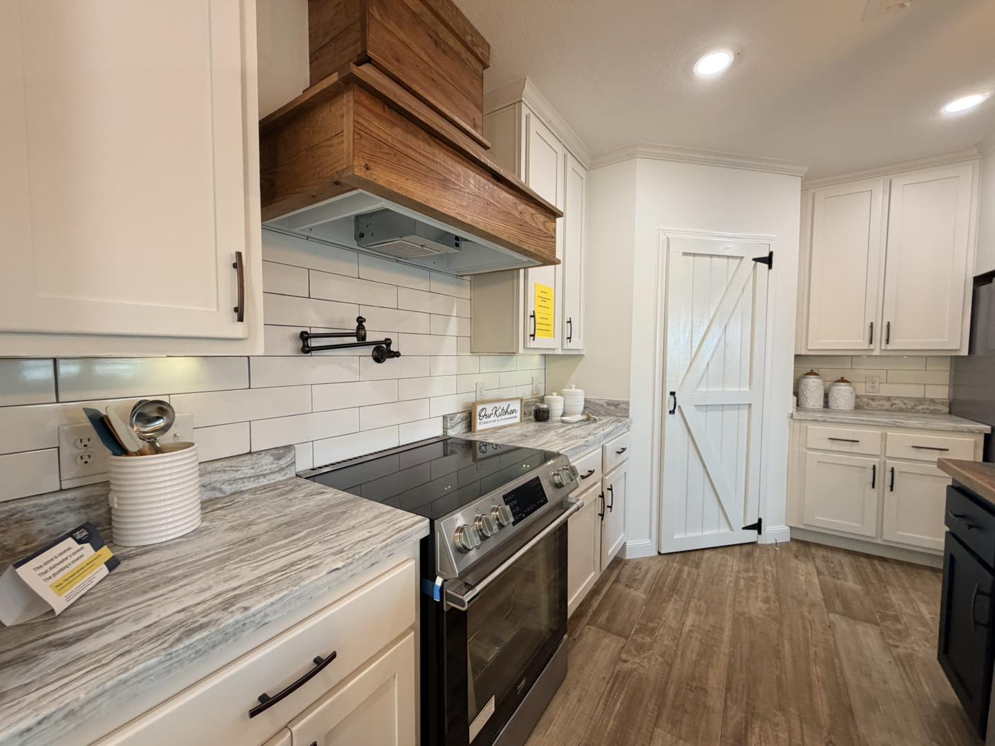 Modern kitchen with white cabinets, wooden range hood, and stainless steel stove. Features white subway tiles, light wood flooring, and a barn door. Cozy and clean ambiance.
