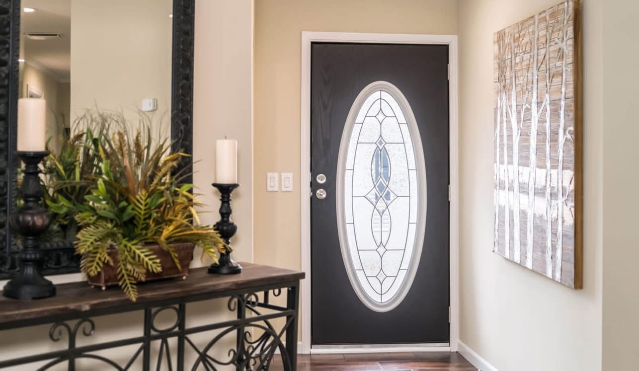 A front entry with a dark door featuring an oval glass design, adjacent wall art, and a console table with candles and plants, creating a welcoming atmosphere.