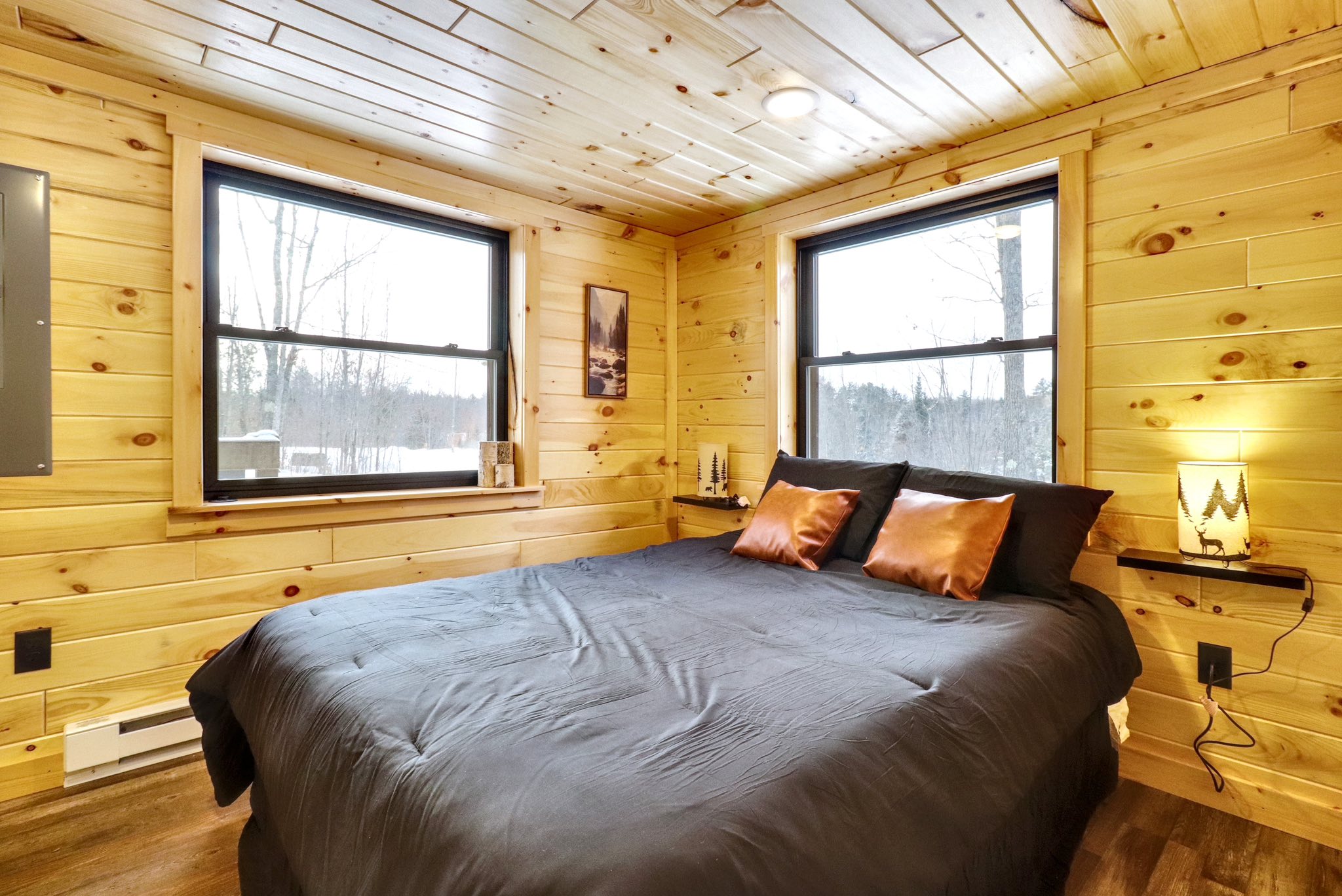 Cozy bedroom with wooden walls and ceiling, featuring two windows, a bed with black bedding, copper pillows, a small nightstand, and soft lighting.