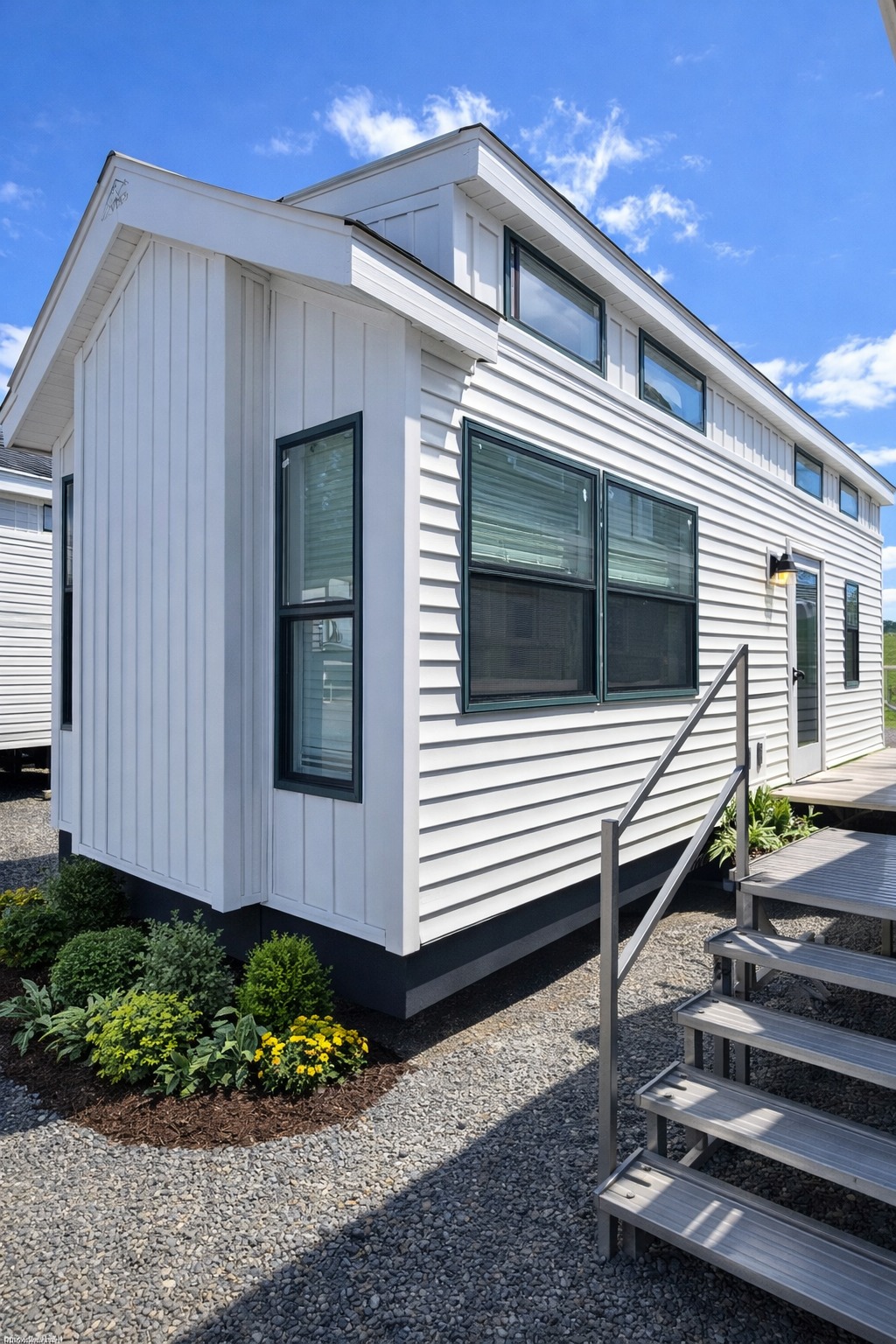 White tiny house with large windows, steps leading to entrance, surrounded by gravel and greenery. The sky is bright blue with scattered clouds.