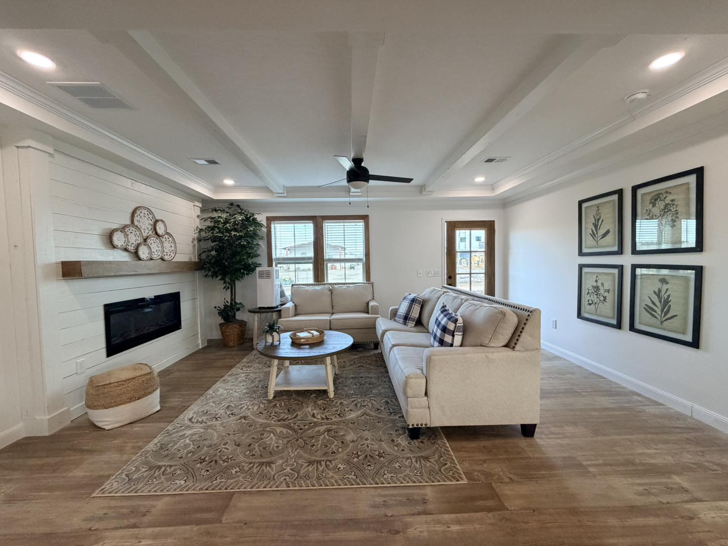 Spacious living room with beige sofas and checkered pillows, wooden coffee table, fireplace, decorative plates, botanical art, and a ceiling fan. Cozy ambiance.