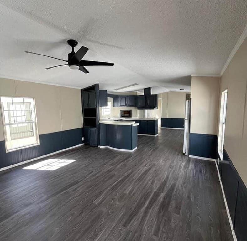Spacious modern kitchen and living area with dark wood floors, beige walls, navy accents, and large windows. Ceiling fan adds a touch of elegance.