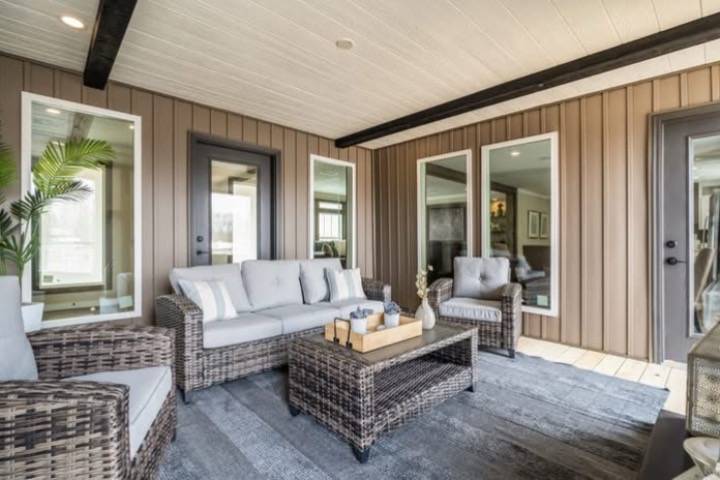 Cozy patio with wicker furniture, gray cushions, and a matching coffee table on a gray rug. Brown paneled walls and large windows create a serene ambiance.