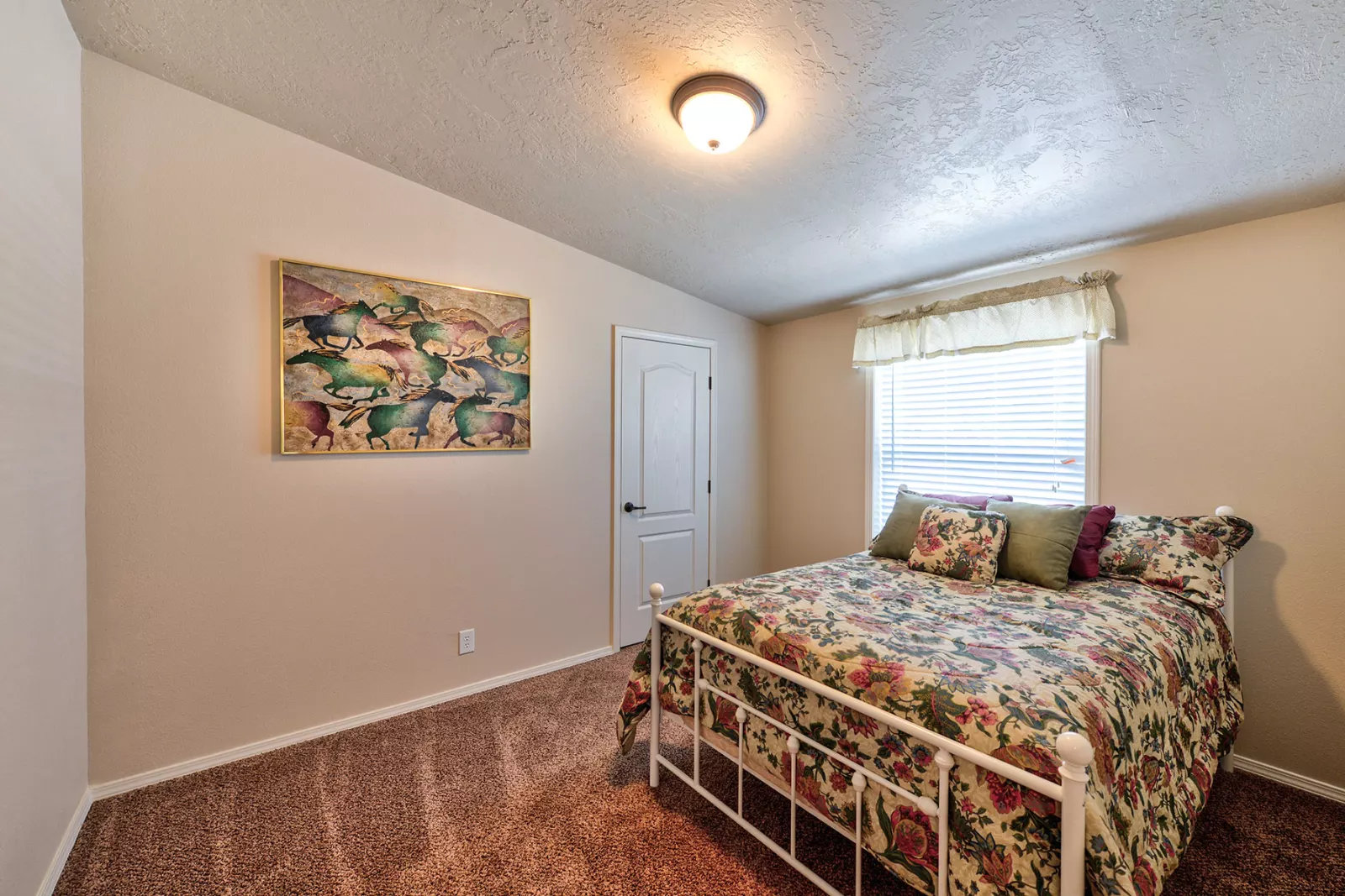 A cozy bedroom with a floral quilt-covered metal bed, decorative pillows, a bright window with a valance, beige walls, a painting, and carpet flooring.