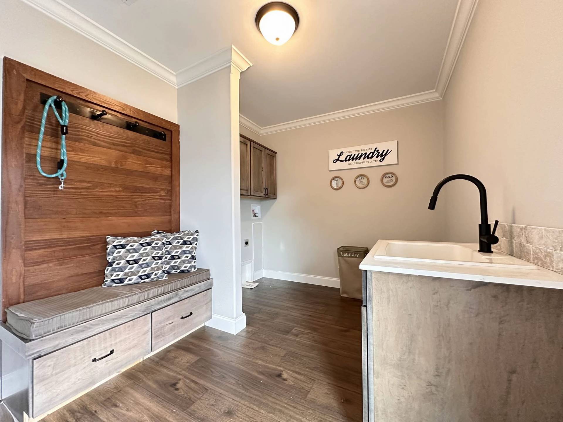 Laundry room with a cozy bench and patterned pillows on the left. A turquoise leash hangs above. A sink with a black faucet is on the right, and a laundry basket under a "Laundry" sign is visible in the neutral-toned space.
