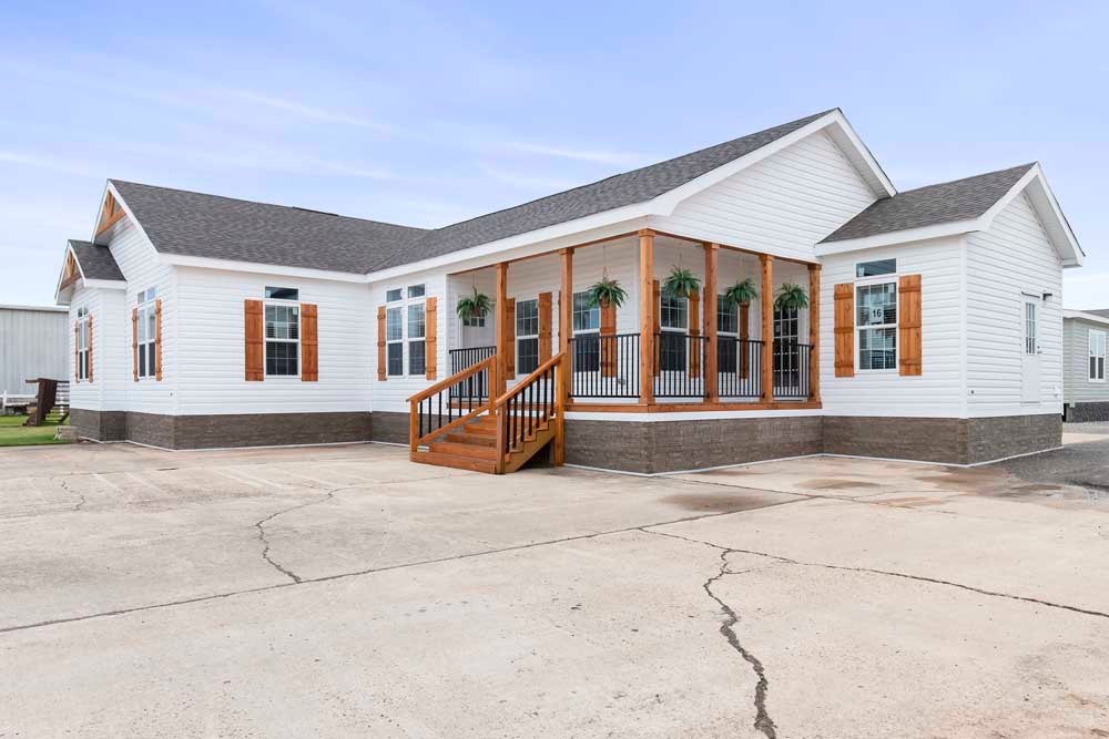 Single-story white modular home with brown shutters and a welcoming porch adorned with hanging plants. The sky is overcast, creating a serene atmosphere.