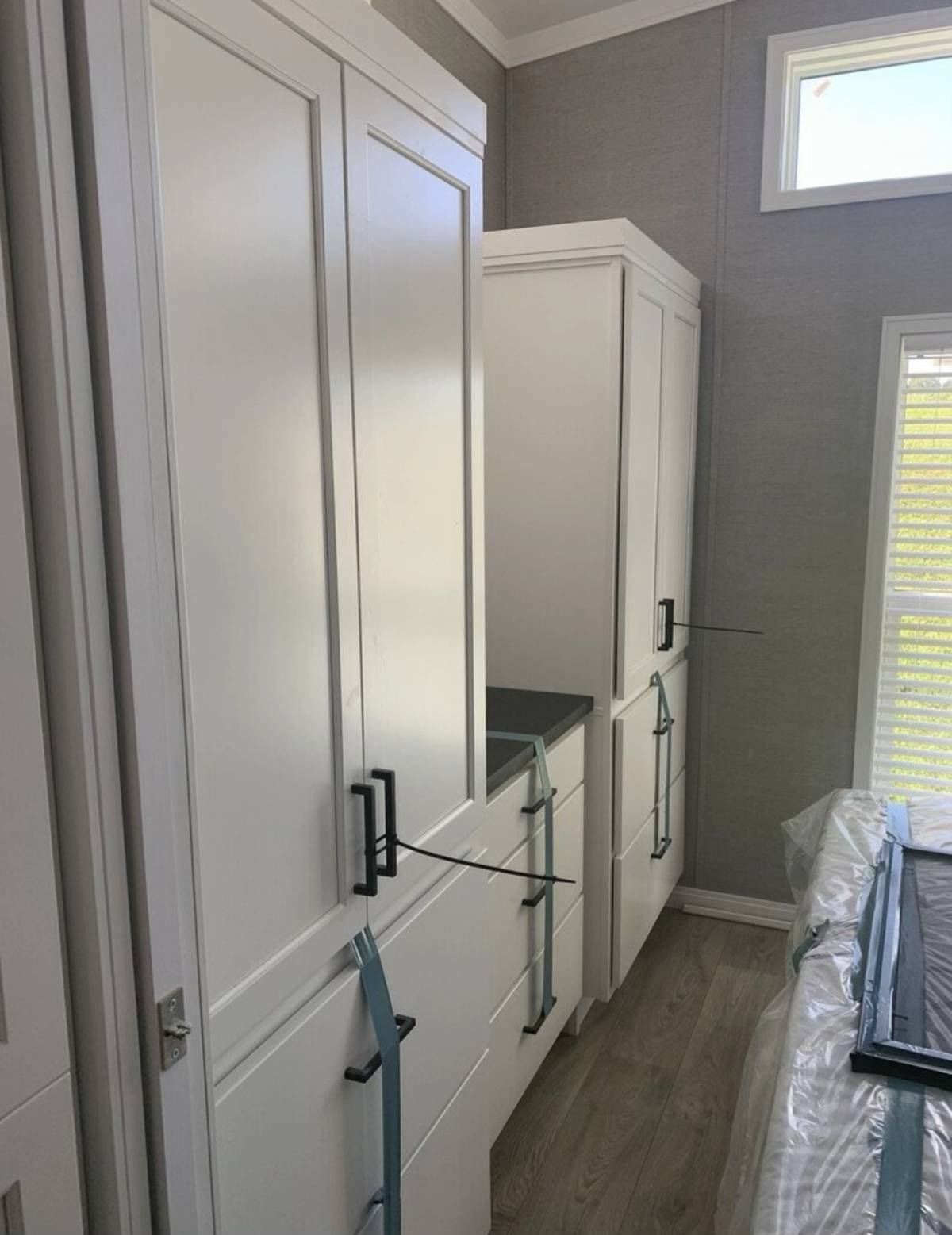 White cabinets with black handles are strapped for safety in a room with gray walls and wood flooring. Soft light from a window creates a calm ambiance.