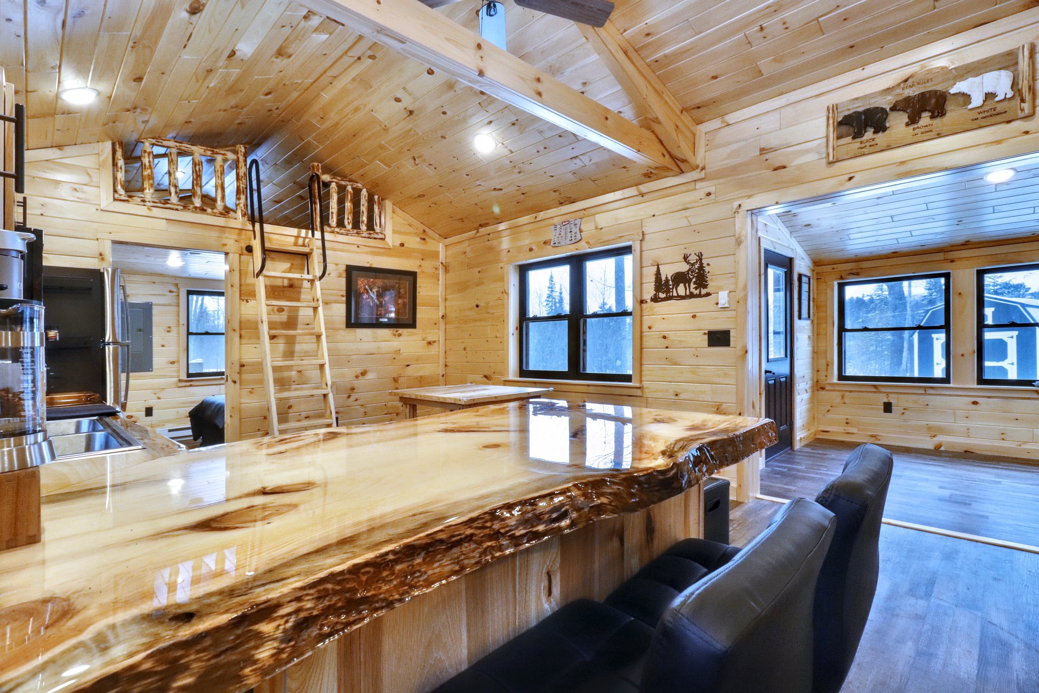 Cozy log cabin interior with wooden walls, a polished wood bar, and black cushioned chairs. A ladder leads to a small loft area, creating a rustic, warm ambiance.
