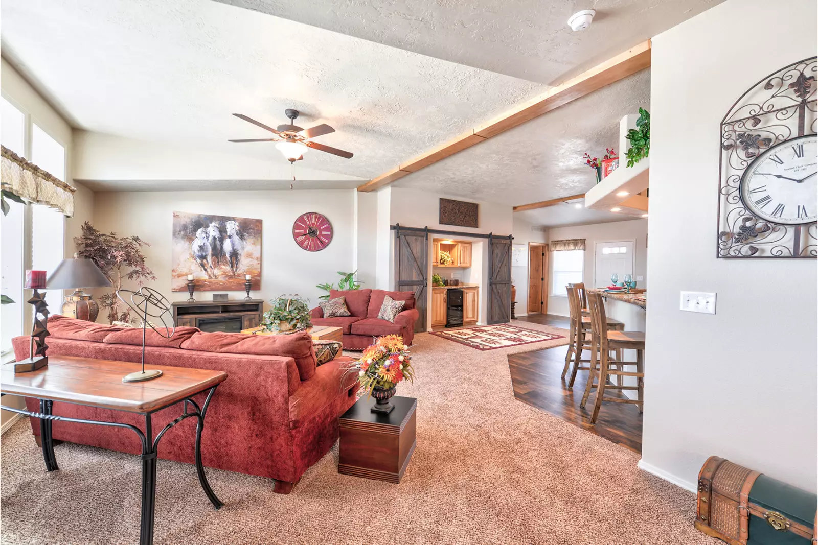 A cozy living room with red sofas, warm lighting, and rustic decor. Features a ceiling fan, wooden beams, wall art, and a large clock, creating an inviting atmosphere.