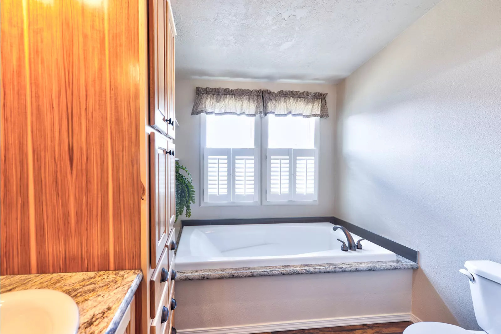 Bright bathroom with a large tub by a window with shutters. Wood cabinets and a granite countertop add warmth. A serene, inviting space.