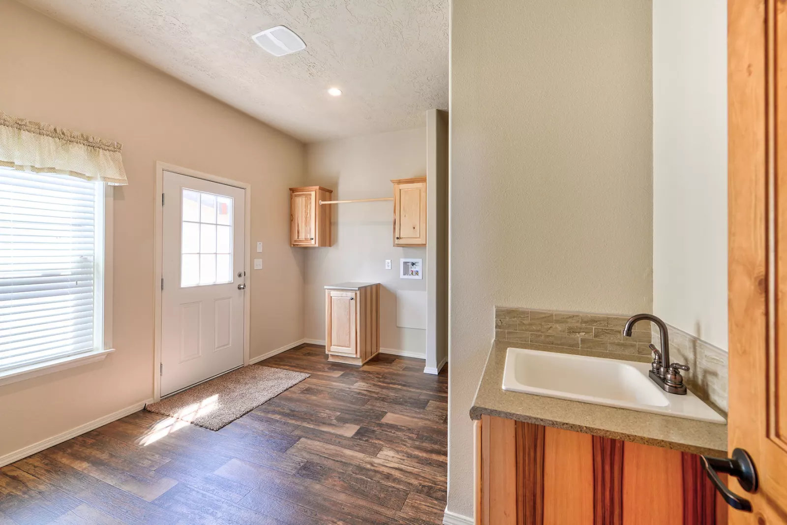 A bright laundry room with wood flooring, featuring cabinets and a countertop with a sink. A door and a window with blinds let in natural light.