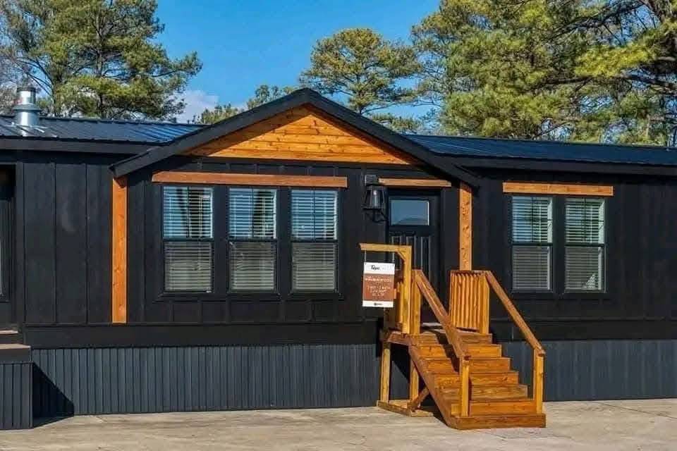 Black modular home with contrasting wooden trim and a small wooden staircase leading to the entrance. Surrounded by tall pine trees under a clear blue sky.