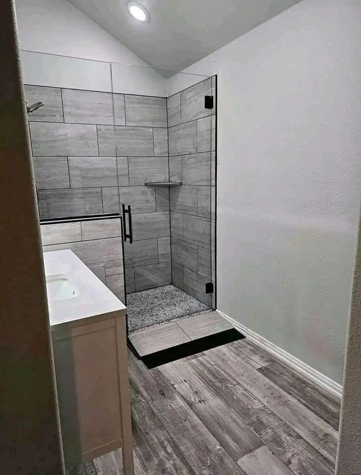 Modern bathroom with a glass-door shower featuring large gray tiles and a small shelf. Light wood flooring and a white vanity create a clean, minimalistic look.