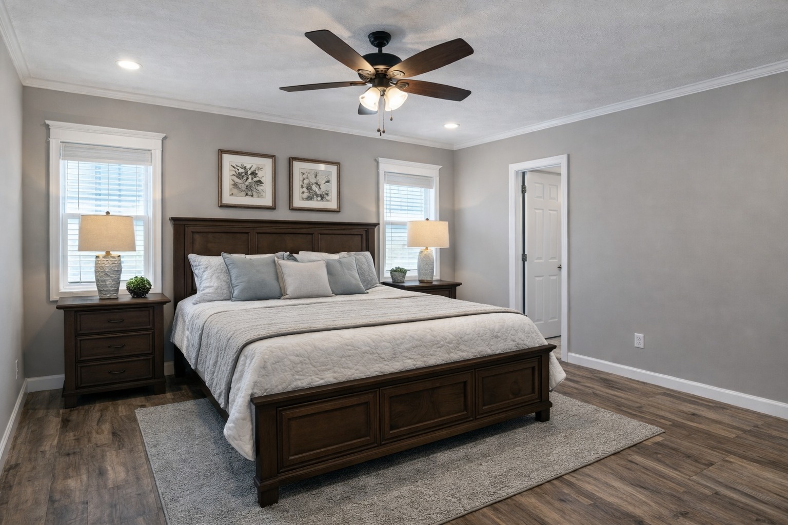 A cozy bedroom features a large wooden bed with a white quilt and gray pillows. Two nightstands with lamps flank the bed. The room is well-lit, with neutral tones and hardwood flooring creating a serene atmosphere.