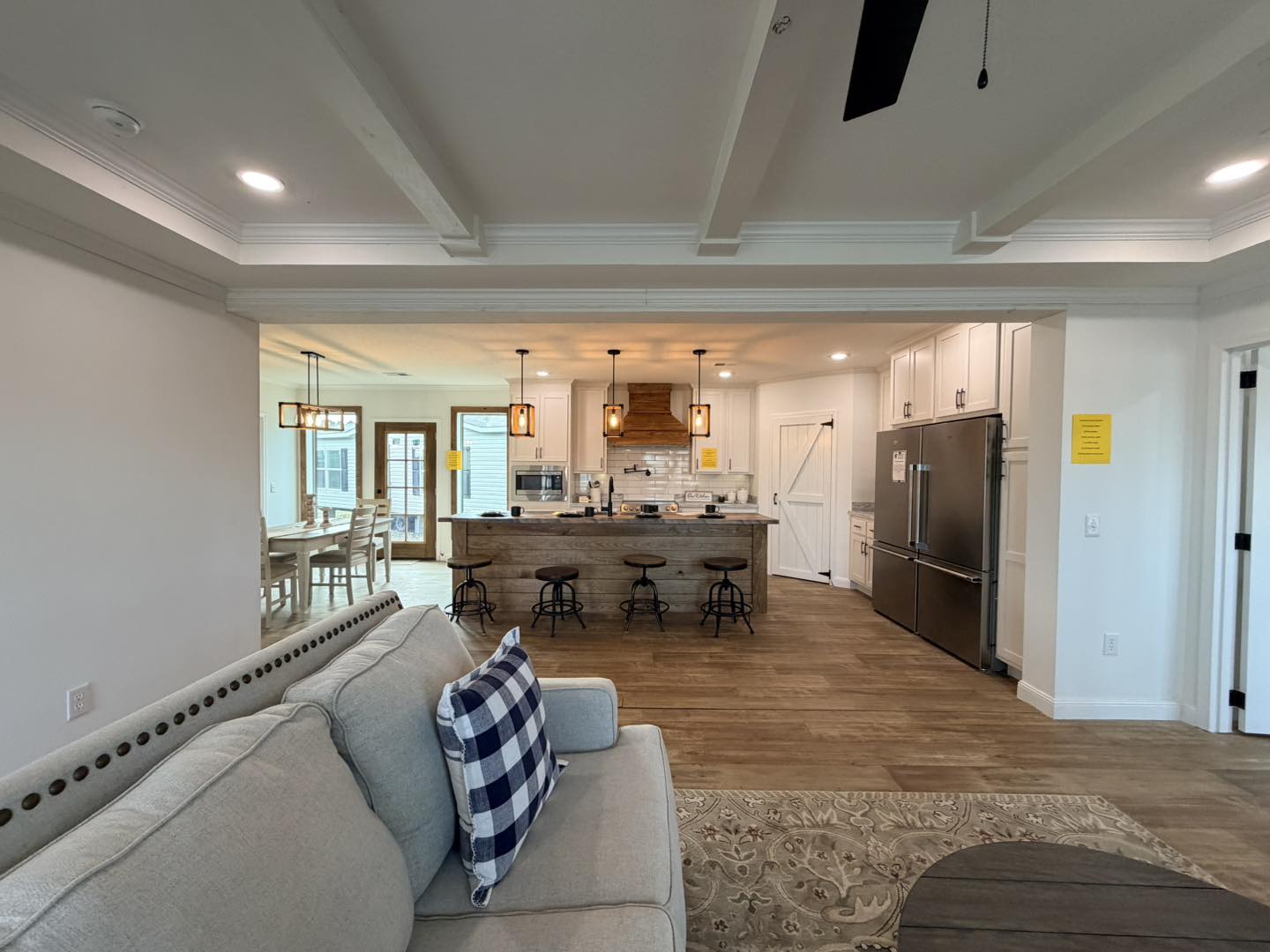Open-concept living space featuring a cozy gray sofa with a checkered pillow, facing a modern kitchen with a wooden island, stools, and pendant lights. Warm, inviting tones.