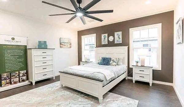 Bright bedroom with modern decor. Features a white bed, matching dresser, nightstands, two windows, wall art, and a large ceiling fan. Calm and inviting.