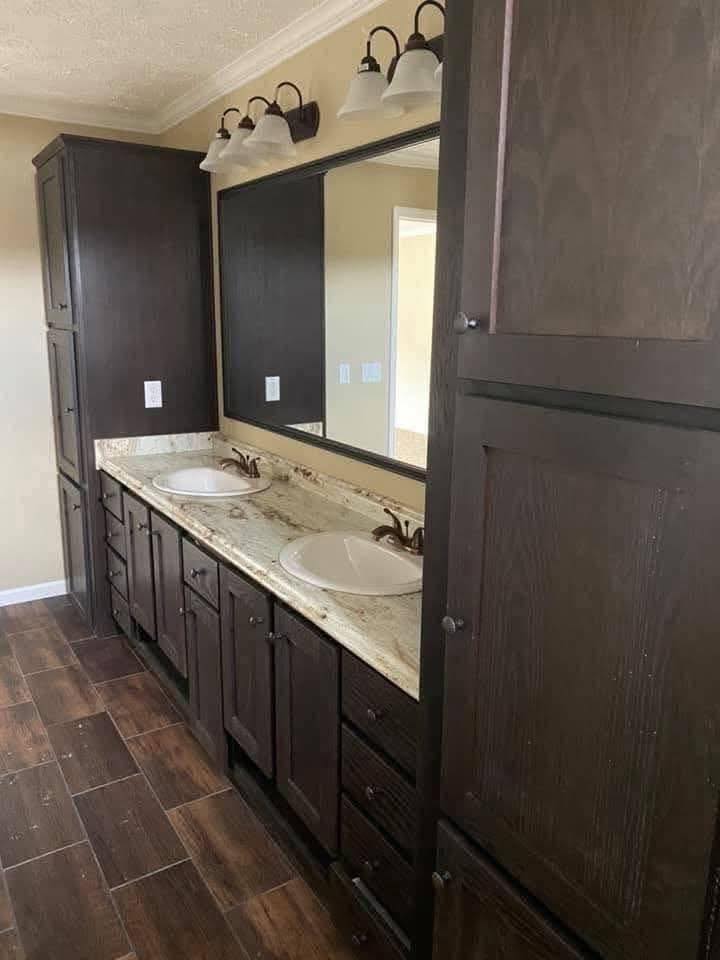 Dark wood bathroom cabinets with two sinks and a large mirror above on a beige wall. Overhead lights illuminate the granite countertop and brown tiled floor.