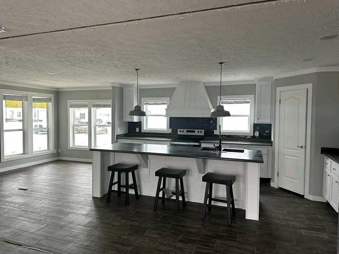 Spacious kitchen with white cabinets, large island, and three dark stools. Pendant lights hang above, contrasting with dark wood floors. Bright light streams through multiple windows, creating an airy feel.