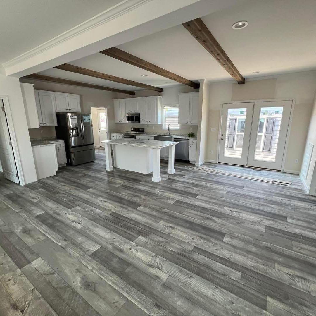 Modern kitchen with rustic charm, featuring gray wood floors, a central island, white cabinets, exposed beams, and double glass doors, promoting a cozy atmosphere.