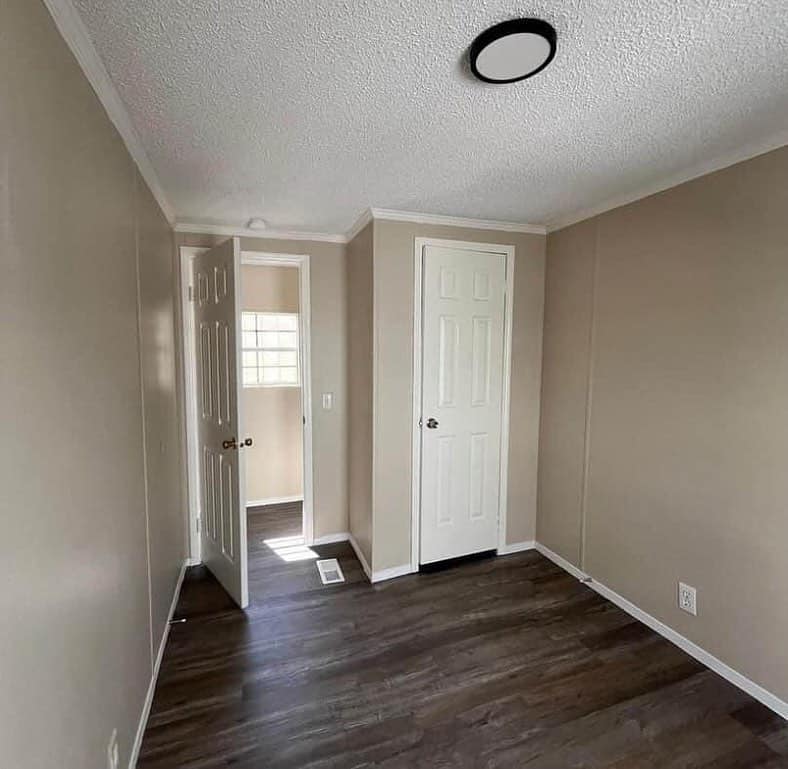 Small room with beige walls and dark wooden flooring. Two closed white doors and an open door showing a lit area. Ceiling has a circular light fixture.