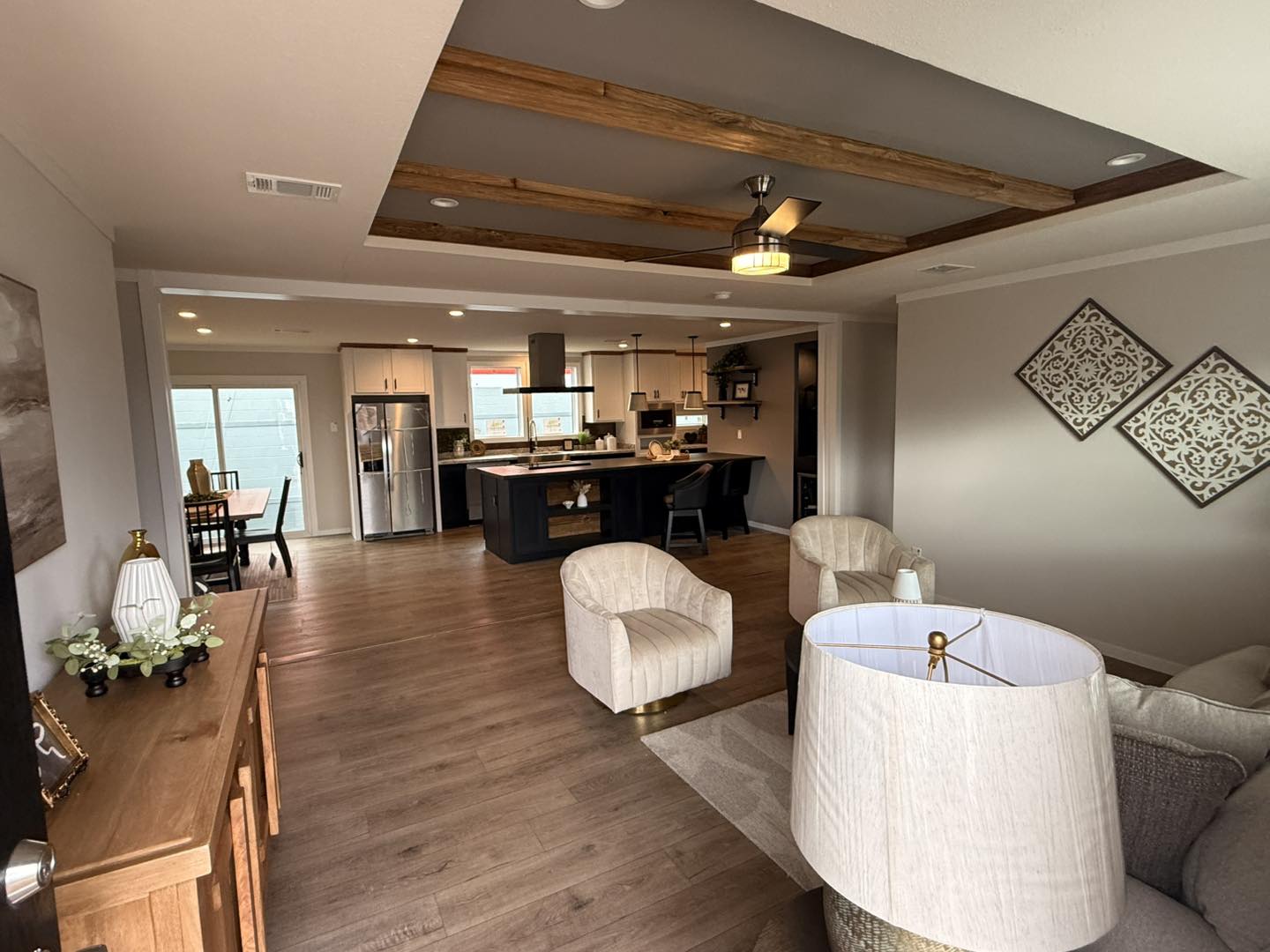 Modern living room leading to an open kitchen and dining area. Features cozy chairs, warm wood flooring, decorative ceiling beams, and soft lighting.