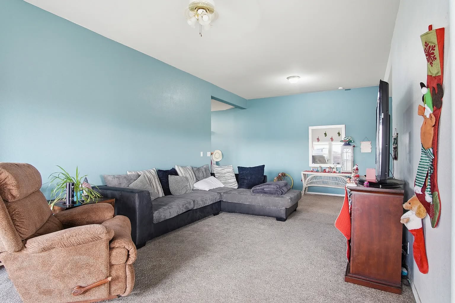 A cozy living room with a blue accent wall, featuring a gray sectional sofa adorned with throw pillows. A brown recliner sits nearby, with a small plant beside it. On the right, a wooden TV stand with a large television is positioned. A festive Christmas stocking hangs on the wall, adding a touch of holiday spirit. The room is well-lit, conveying a welcoming and relaxed atmosphere.
