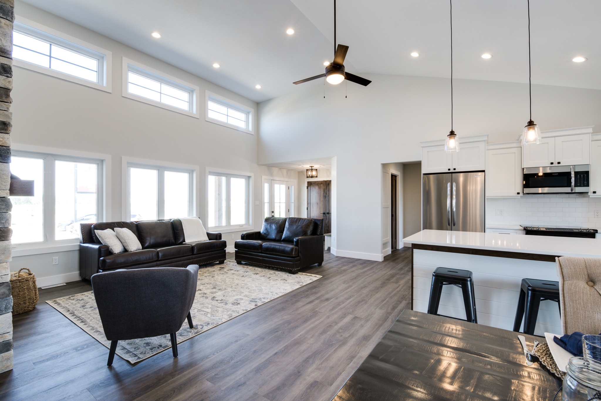 Spacious modern living room and kitchen with high ceilings, featuring large windows, dark leather sofas, wood floors, and sleek kitchen island with lights.