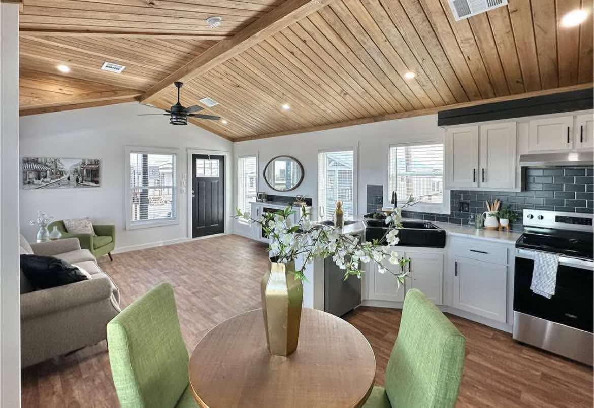 Cozy open-concept room with wooden ceiling, modern kitchen, green chairs, round table, and elegant decor; bright, inviting ambiance.